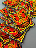 A pile of die-cut stickers featuring a stylized red, orange, and yellow skull face with black outlines and holographic glitter borders, set against a neutral gray background.