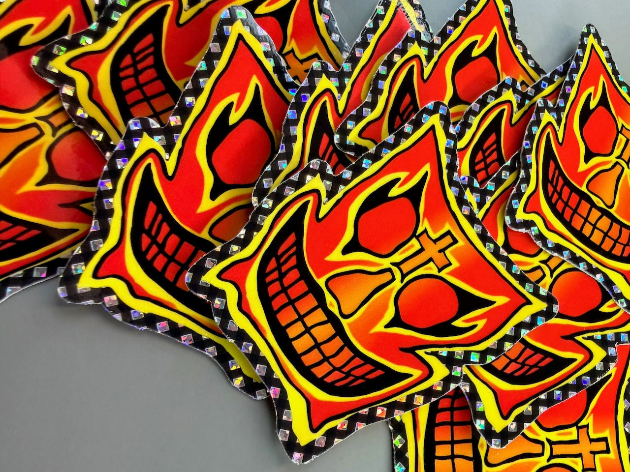 A pile of die-cut stickers featuring a stylized red, orange, and yellow skull face with black outlines and holographic glitter borders, set against a neutral gray background.