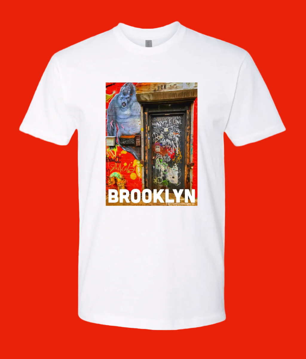 A white short-sleeve crewneck t-shirt featuring a colorful graphic print of a Brooklyn street scene with a blue gorilla and the word 'BROOKLYN' in bold white letters.