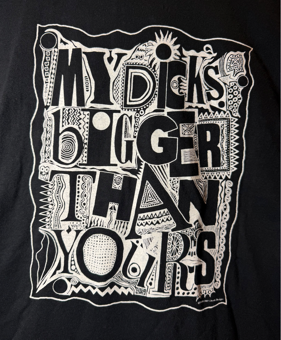 A close-up of a black graphic t-shirt featuring a large white hand-drawn style illustration with the text 'MY DICKS BIGGER THAN YOURS' surrounded by doodles and abstract shapes.