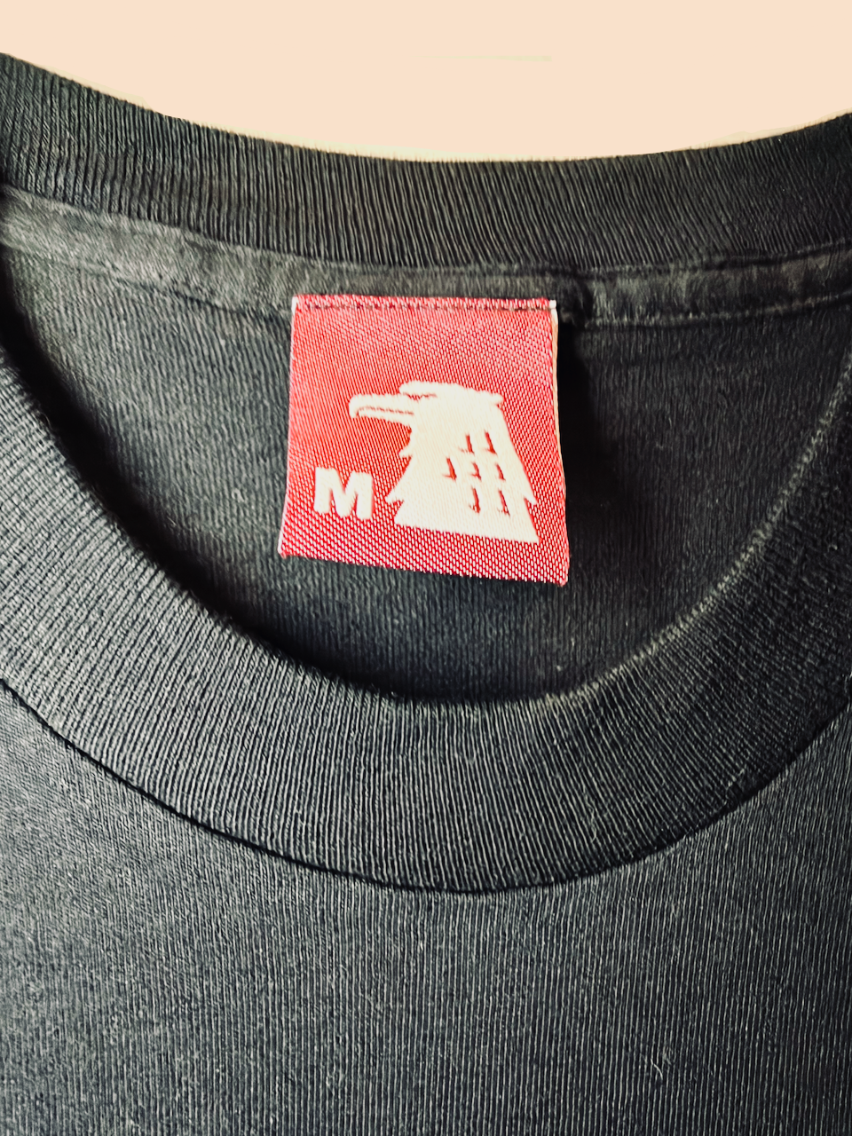 Close-up of a black crewneck t-shirt showing a red neck label with a white bird logo and the letter 'M'.