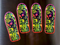 Four skateboard decks arranged in a row on a dark metallic surface, featuring a vibrant neon green, yellow, pink, and black psychedelic skull and crossbones design.