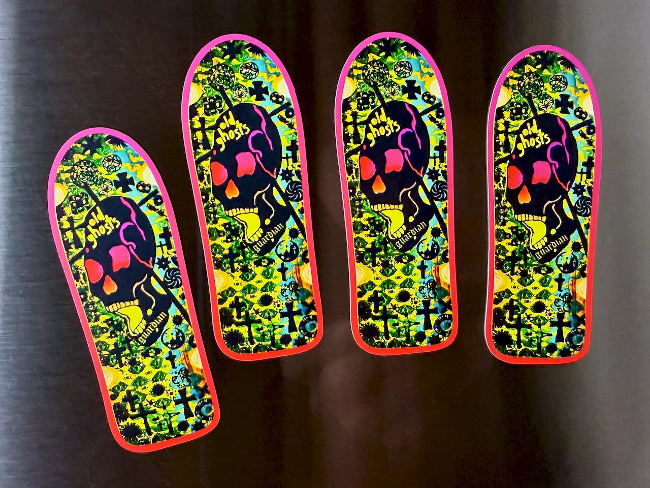Four skateboard decks arranged in a row on a dark metallic surface, featuring a vibrant neon green, yellow, pink, and black psychedelic skull and crossbones design.