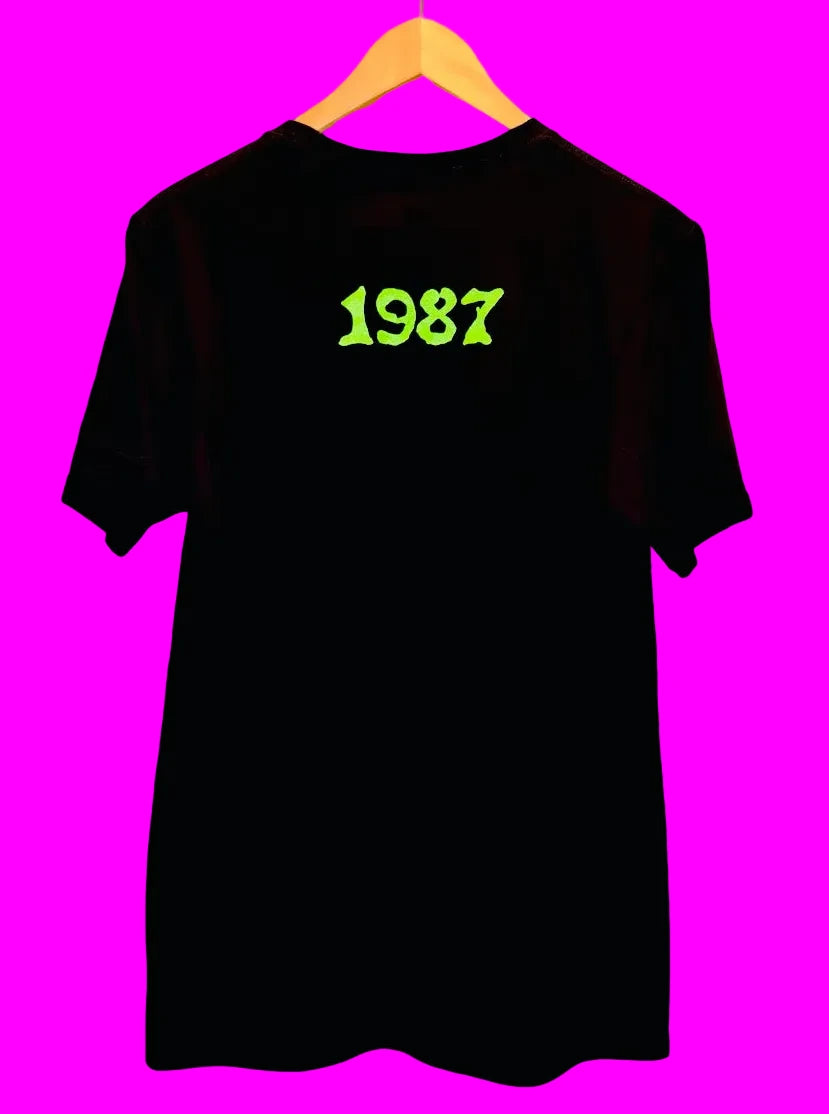 A black short-sleeve t-shirt hanging on a wooden hanger against a PINK background, featuring the year '1987' printed in a bright neon green font on the chest.