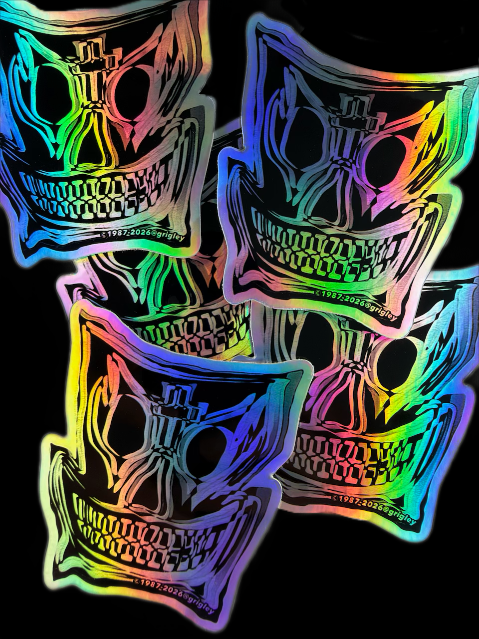 Four die-cut vinyl stickers featuring a stylized skull with a cross on its forehead, rendered in a vibrant rainbow holographic foil finish against a solid black background.