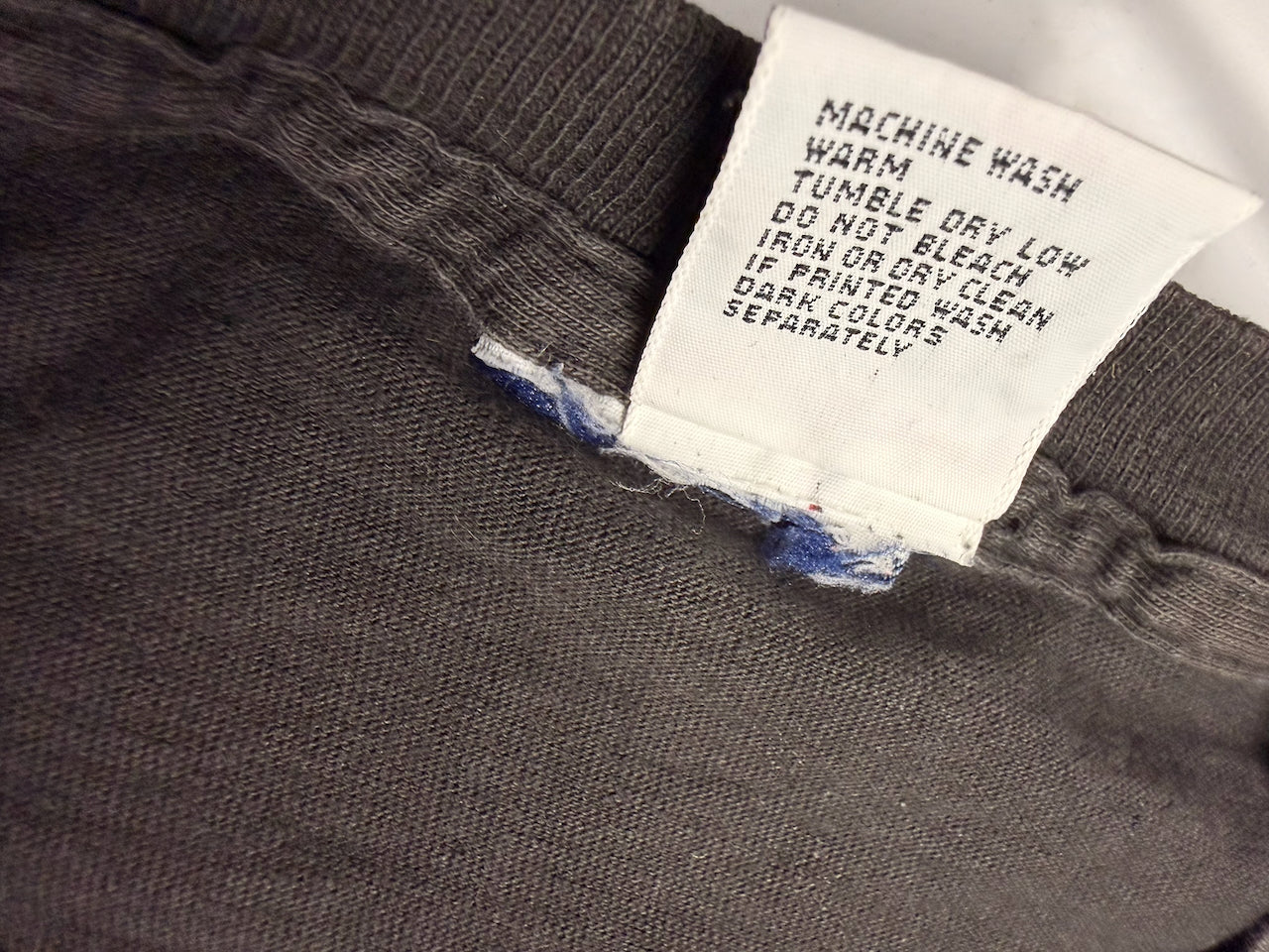 Close-up of a dark gray knit garment with a white sewn-in care label reading 'MACHINE WASH WARM TUMBLE DRY LOW DO NOT BLEACH IRON OR DRY CLEAN IF PRINTED WASH DARK COLORS SEPARATELY'.