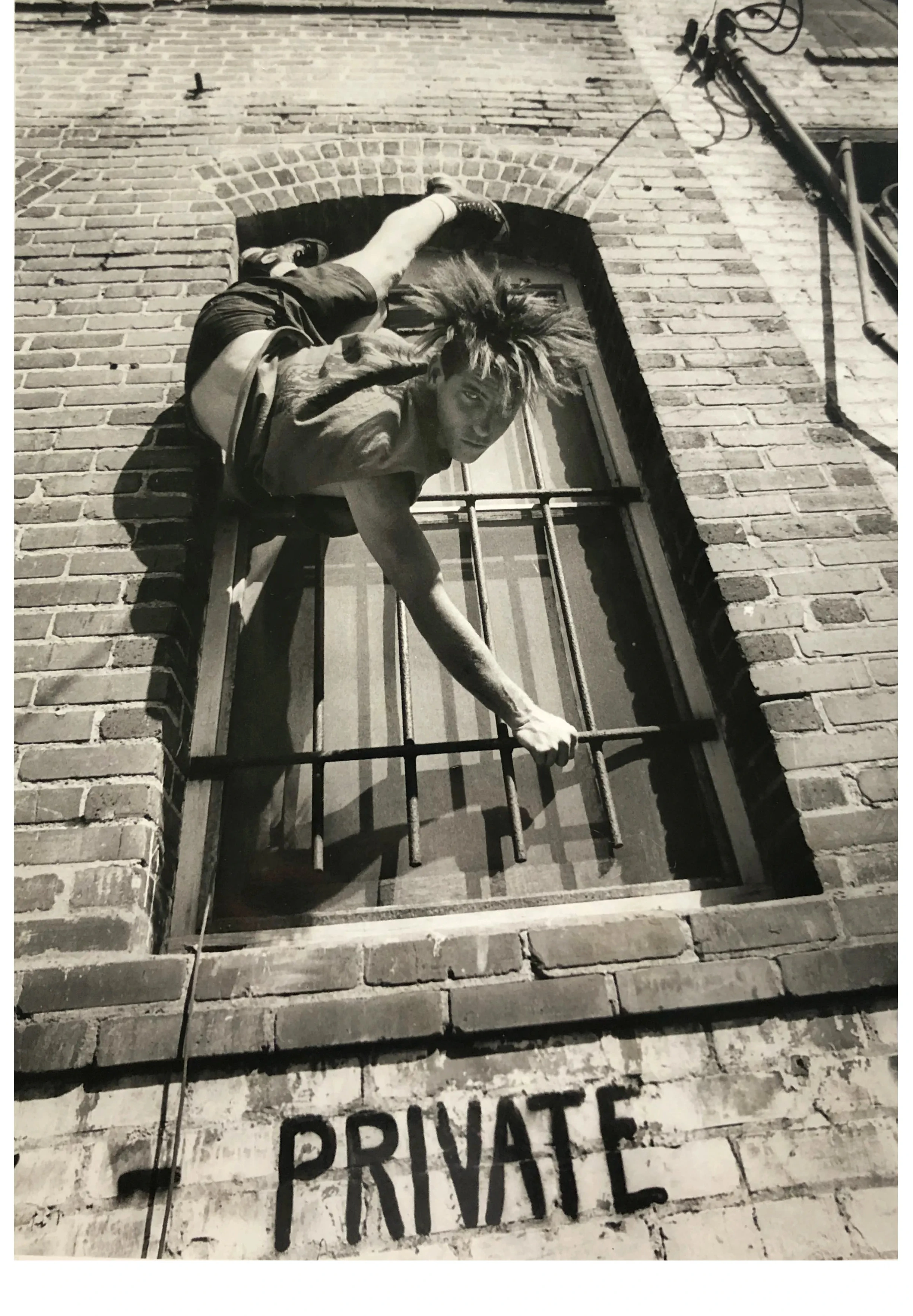 A black and white art photograph of a person climbing onto a brick building window with metal bars, with the word 'PRIVATE' painted on the brick below.