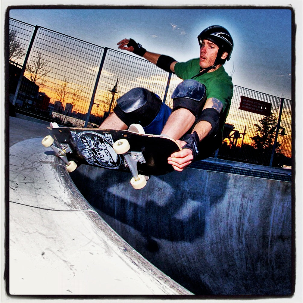 A skateboarder in a green shirt and black helmet rides a skateboard along the curved edge of a concrete skate bowl at sunset, with a metal fence and trees in the background.