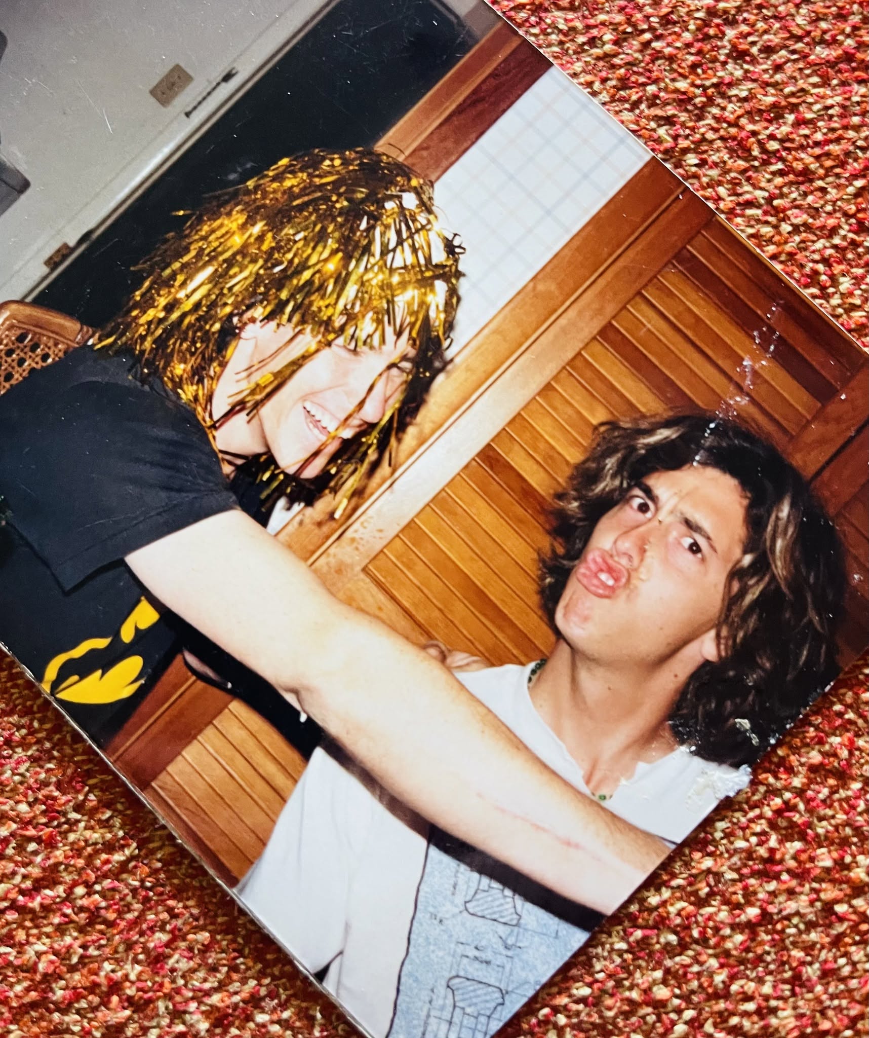 Two people sitting on a couch with a wooden floor and textured red and brown background.