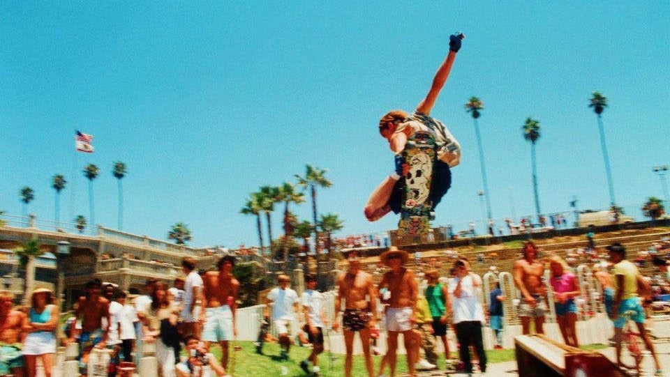 Person performing a skateboard trick in a sunny outdoor setting with palm trees and a crowd.