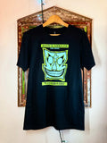 A black short-sleeve t-shirt hanging on a wooden hanger against a wall in front of a decorative picture frame. The shirt features a large lime green graphic of John Gotti's iconic skull face with the text 'JOHN GOTTI FOR HONOR' and 'VISITOR 1987'.