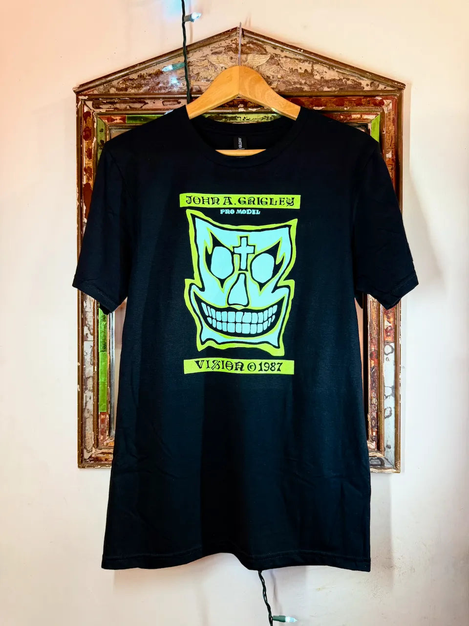 A black short-sleeve t-shirt hanging on a wooden hanger against a wall in front of a decorative picture frame. The shirt features a large lime green graphic of John Gotti's iconic skull face with the text 'JOHN GOTTI FOR HONOR' and 'VISITOR 1987'.