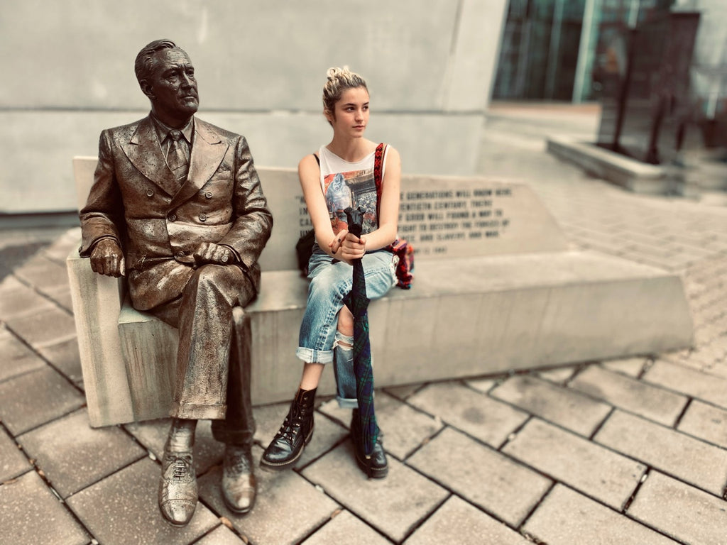 A woman sits on a stone bench next to a bronze statue of a suited figure, outdoors on a paved plaza with a modern building in the background.