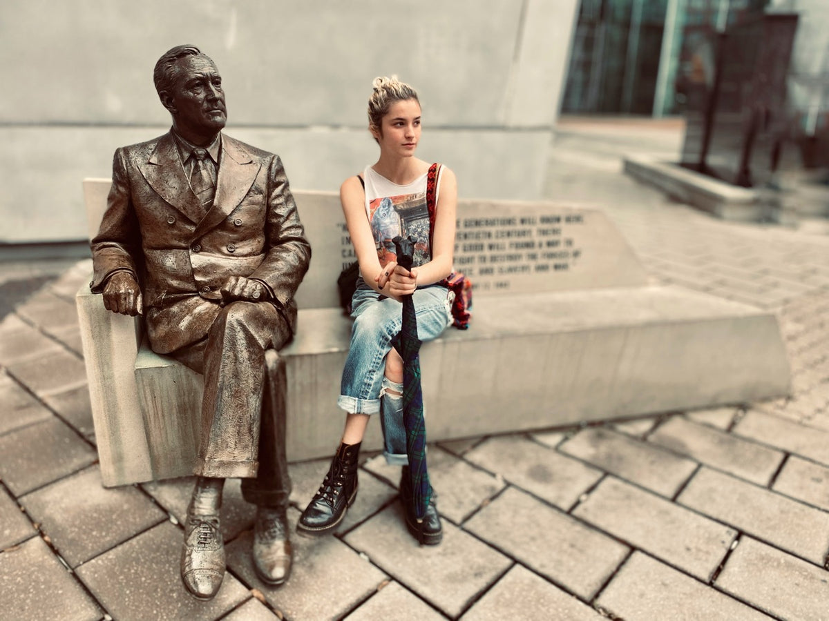 A woman sits on a stone bench next to a bronze statue of a suited figure, outdoors on a paved plaza with a modern building in the background.