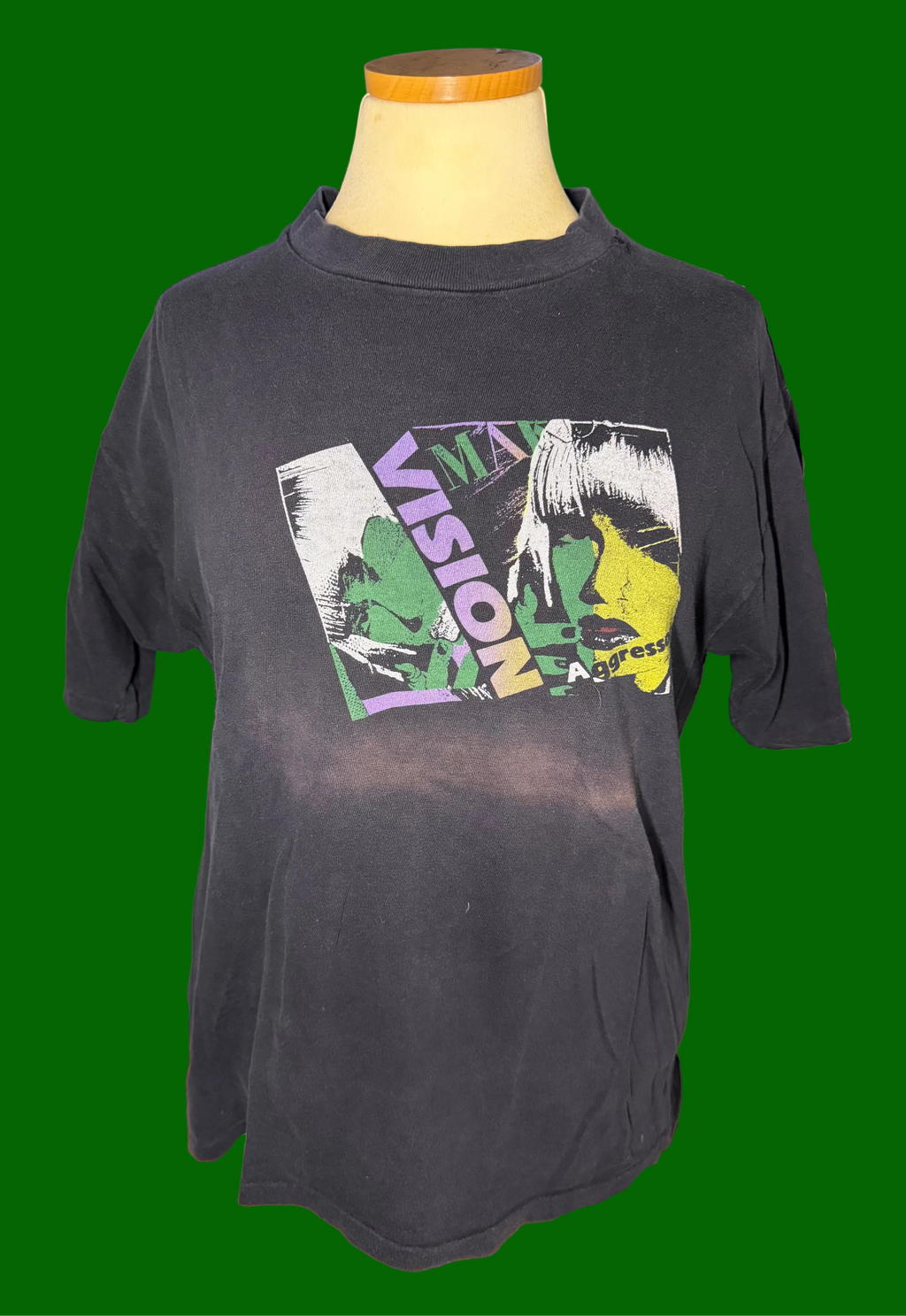 A faded black graphic t-shirt displayed on a mannequin torso on a green background. The shirt features a large, colorful front graphic with the word 'VISION' and a stylized illustration in green, yellow, and white.