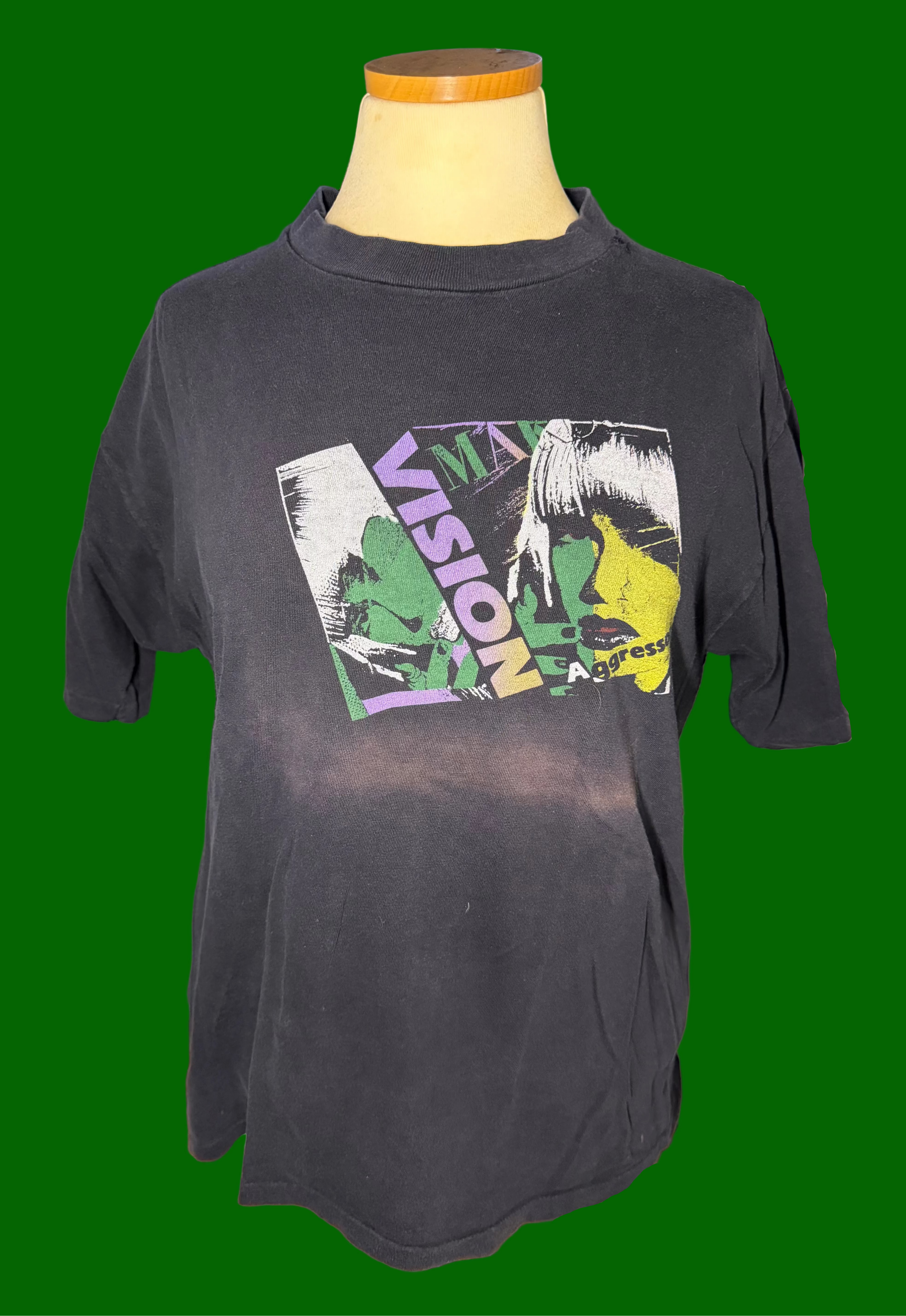 A faded black graphic t-shirt displayed on a mannequin torso on a green background. The shirt features a large, colorful front graphic with the word 'VISION' and a stylized illustration in green, yellow, and white.